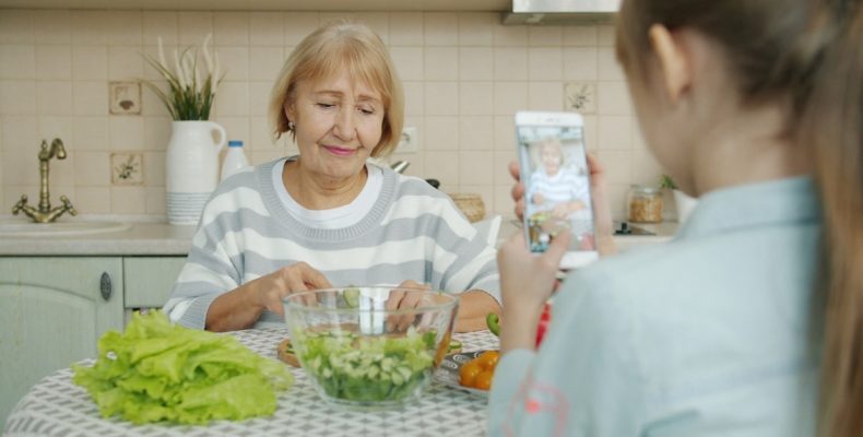 Little girl is taking pictures of granny who is cooking salad then posing for camera with cucumber slices having fun, child is using modern smartphone camera.